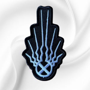 May include: A black embroidered patch featuring a light blue design of a central circle with an X inside, from which five vertical, wavy lines extend upward in a fan-like arrangement, creating a symmetrical, stylized emblem shape.
