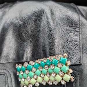 Peut inclure: Broche décorative avec un motif tissé de pierres rectangulaires vertes et vert clair, entrecoupées de cristaux ronds et transparents. La broche a des accents en métal doré, posée sur une surface en cuir noir.