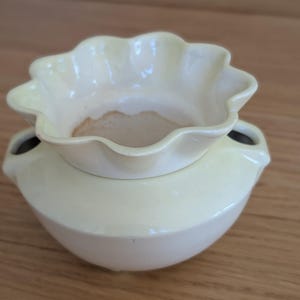 May include: A pale yellow ceramic oil burner with a scalloped, flower-shaped top bowl and a rounded base. The bowl features a smooth, glossy finish with a light brown residue at the bottom. Two small circular openings are visible on the sides.