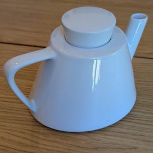 Peut inclure: Une théière en céramique blanche avec une finition lisse et brillante, dotée d'une base conique large, d'un couvercle circulaire plat, d'une anse incurvée sur le côté gauche et d'un bec verseur droit et incliné sur le côté droit.