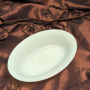 May include: An oval, cream-coloured serving dish. The dish is empty and has a smooth, glossy surface. The background is a brown fabric with a rose pattern.