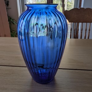 May include: A striking blue glass vase with vertical ribbed detailing. The vase has a rounded body and a slightly flared rim. The glass reflects light. The vase is on a wooden table. The vase is approximately 25 cm tall.