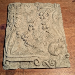 May include: A rectangular, weathered, light-colored stone relief with a botanical design. The relief features a branch with berries, leaves, and decorative scrollwork. The stone has a rough texture and is set against a wooden surface.