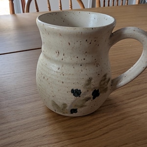 Peut inclure: Une tasse en céramique beige mouchetée avec une anse incurvée. La tasse présente un motif peint à la main de baies bleu foncé et de feuilles vertes. La tasse est posée sur une surface en bois.
