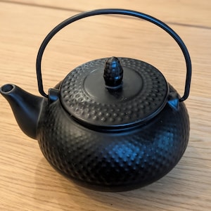 May include: A black cast iron teapot with a textured hobnail pattern on the body. The teapot features a curved spout, a matching removable lid with a small knob handle, and a thin, arched metal handle that extends over the top of the vessel.
