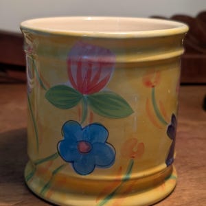 May include: A yellow ceramic jar decorated with hand-painted flowers. The flowers are blue, red, and purple, with green leaves and stems. The jar has a cream interior and a rounded base.
