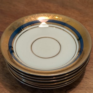 Peut inclure: Une pile de six soucoupes blanches avec des bords dorés et une bande bleue. Les soucoupes ont un cercle intérieur doré et sont empilées sur une surface en bois. Le design est élégant et classique.