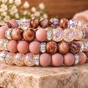 May include: A stack of three beaded bracelets featuring a mix of matte dusty rose beads, brown marbled beads, clear crackle glass beads with iridescent finishes, and silver-toned spacers encrusted with small clear crystals.