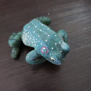 May include: A handcrafted frog figurine in shades of teal and green, adorned with white speckles. The frog has red-rimmed eyes with white dots and yellow accents. The figurine is placed on a dark wooden surface.