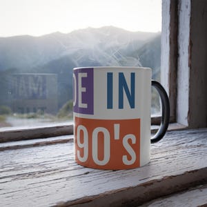 May include: A white ceramic mug with the text "MADE IN 90's" in purple, blue, and orange. The mug has a black handle and is steaming. The mug is sitting on a white, weathered wooden surface.