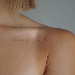 Może przedstawiać: Zbliżenie na ramię i górną część klatki piersiowej osoby z tatuażem "amor fati" w kursywie. Skóra jest jasna, z małym czerwonym znakiem. Tło jest gładkie, neutralnie szare.