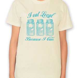 May include: A white t-shirt with a blue graphic that says "I eat local because I can". The graphic features three blue mason jars with the text "Ball Perfect Mason" on each jar.