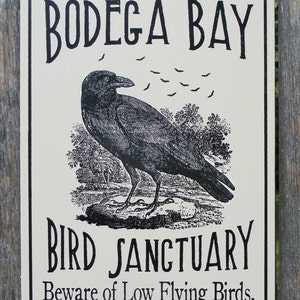May include: A black and white sign with the text "Bodega Bay Bird Sanctuary" and the warning "Beware of Low Flying Birds". The sign features a black crow with a flock of bats flying above it.