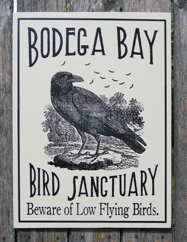 May include: A black and white sign with the text "Bodega Bay Bird Sanctuary" and the warning "Beware of Low Flying Birds". The sign features a black crow with a flock of bats flying above it.