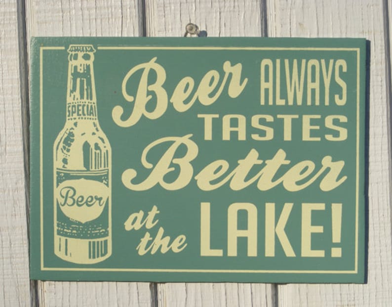 May include: Green wooden sign with white text that reads "Beer Always Tastes Better at the Lake!"  A vintage-style illustration of a beer bottle is on the left side of the sign.