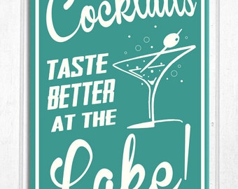 Op de afbeelding: Teal en wit bord met de tekst "Cocktails smaken beter aan het meer!". Een martini glas illustratie is afgebeeld. Het bord is wit omlijst en geplaatst tegen een witte houten achtergrond.