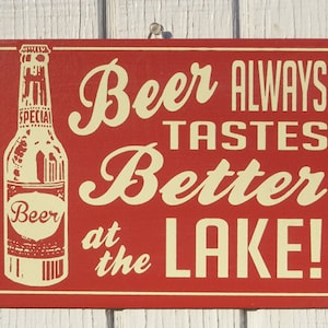 May include: A red wooden sign with white lettering that reads "Beer Always Tastes Better at the Lake!"  A vintage-style illustration of a beer bottle with a label that reads "Beer" is on the left side of the sign.
