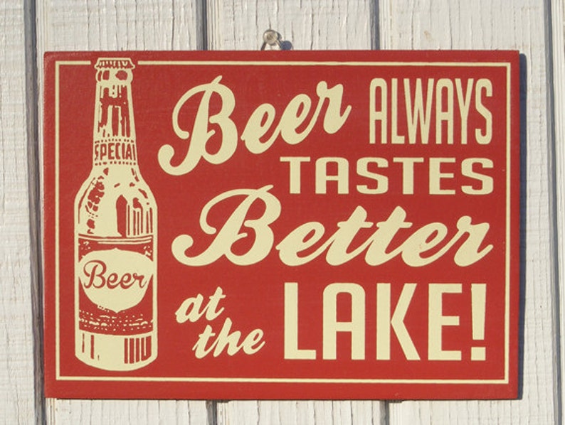 May include: A red wooden sign with white lettering that reads "Beer Always Tastes Better at the Lake!"  A vintage-style illustration of a beer bottle with a label that reads "Beer" is on the left side of the sign.