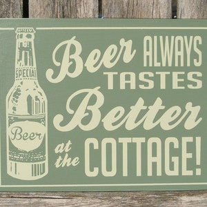 May include: Green wooden sign with white text that reads "Beer Always Tastes Better at the Cottage!"  A vintage-style illustration of a beer bottle is on the left side of the sign.