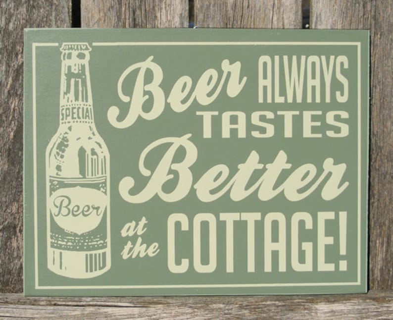 May include: Green wooden sign with white text that reads "Beer Always Tastes Better at the Cottage!"  A vintage-style illustration of a beer bottle is on the left side of the sign.
