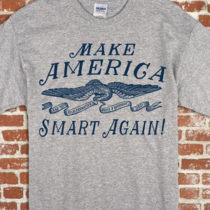 May include: A grey t-shirt with the text "MAKE AMERICA SMART AGAIN!" in blue lettering. The text is surrounded by a banner with the text "SAY NO TO ALTERNATIVE FACTS & OUTRIGHT LIES!" in smaller blue lettering. A blue eagle with wings spread is above the banner.