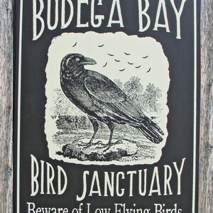 May include: A black wooden sign with white text that reads "Bodega Bay Bird Sanctuary Beware of Low Flying Birds." The sign features a black and white illustration of a crow with a flock of bats flying overhead.