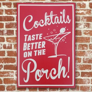 Op de afbeelding: Een rood houten bord met witte tekst die "Cocktails Taste Better On The Porch!" luidt, met een witte illustratie van een martini-glas met een olijf.