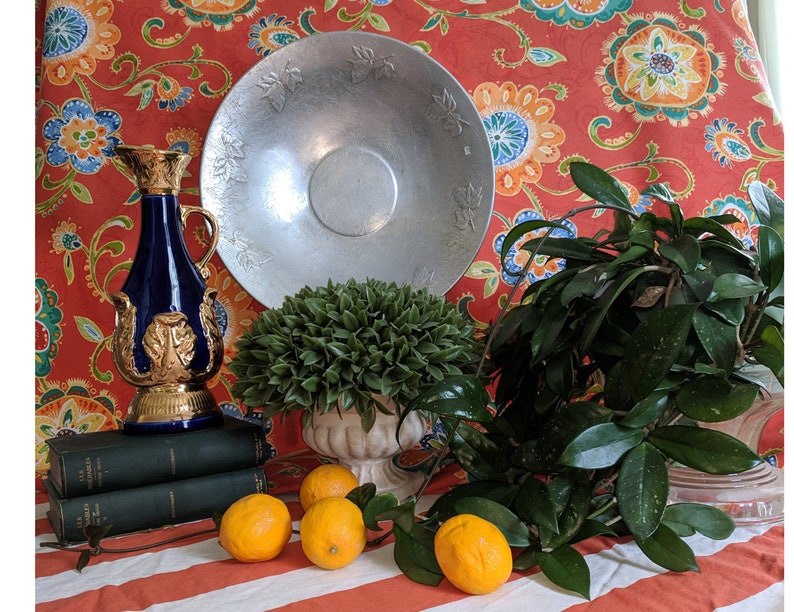 Puede incluir: Un bodeg&oacute;n con una botella decorativa azul y dorada, una fuente de servir plateada, una planta verde en maceta y cuatro limones sobre un mantel a rayas rojas y blancas.