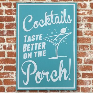 Op de afbeelding: Een tealblauw bord met witte tekst die "Cocktails Taste Better On The Porch!" zegt. Een wit martini-glas met een witte olijf op een stokje is afgebeeld op het bord.