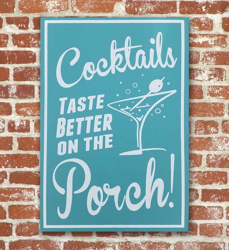 Op de afbeelding: Een tealblauw bord met witte tekst die "Cocktails Taste Better On The Porch!" zegt. Een wit martini-glas met een witte olijf op een stokje is afgebeeld op het bord.
