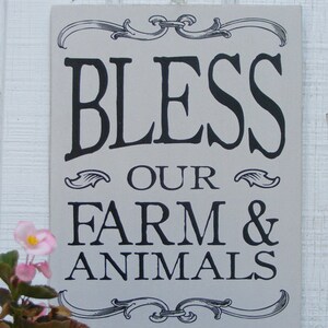 May include: A rectangular, light gray sign with black text that reads "BLESS OUR FARM & ANIMALS." The sign has decorative scrollwork at the top and bottom. Pink flowers are visible in the lower left corner.