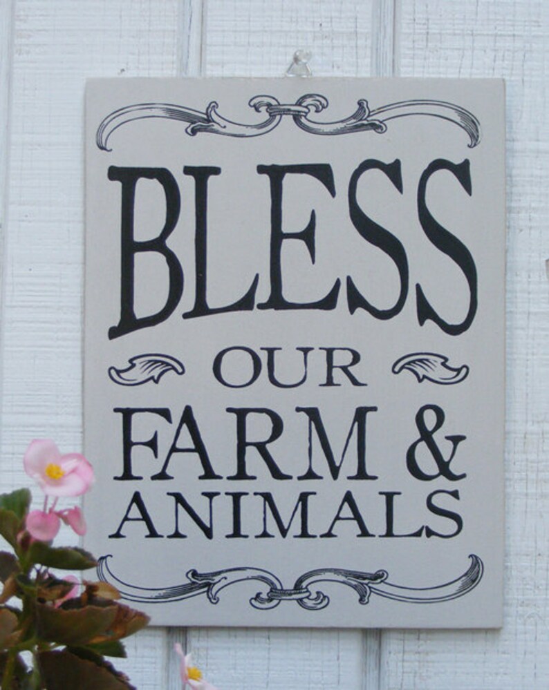 May include: A rectangular, light gray sign with black text that reads "BLESS OUR FARM & ANIMALS." The sign has decorative scrollwork at the top and bottom. Pink flowers are visible in the lower left corner.