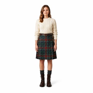 May include: A cream cable knit jumper worn with a knee-length green tartan kilt featuring a brown leather belt with a buckle and side straps. The outfit is completed with dark grey ribbed socks and brown leather lace-up boots.