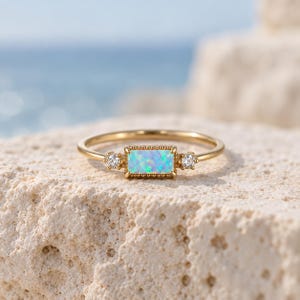 May include: A gold ring featuring a rectangular baguette-cut opal center stone with a vibrant blue and green play-of-color, flanked by two small round clear stones on either side, set in a delicate gold band with a beaded metal border.