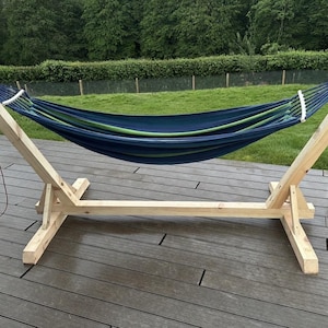 Puede incluir: Una hamaca a rayas azules y verdes suspendida de un marco de madera natural. La hamaca está sostenida por un soporte de madera resistente con un diseño de travesaño. El soporte es de madera clara y está colocado sobre una terraza de madera con césped verde al fondo.