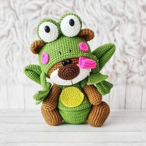 May include: A handmade crochet teddy bear wearing a green frog hat with large white and black eyes, pink cheek spots, and a pink tongue. The bear has brown arms and legs, a white muzzle, and a yellow belly patch. The background is a soft, white, textured surface.