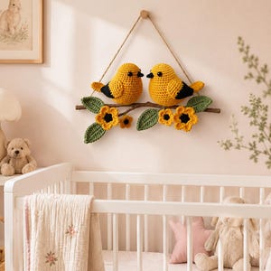 May include: A wall hanging featuring two crocheted yellow birds with black wing accents perched on a brown branch. The branch is decorated with crocheted green leaves and yellow flowers with dark centers. It hangs from a simple tan twine string.