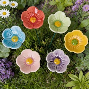 May include: A set of five ceramic flower garden stakes featuring glossy petals in orange, light green, blue, pink, and purple. Each flower has a raised yellow center and is attached to a thin green metal stem, arranged in a cluster.