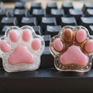 May include: Two paw-shaped resin charms featuring raised pink pads. The left charm has a clear, glitter-infused base, while the right charm features a brown, translucent base. Both items have a glossy, smooth finish with visible air bubbles.