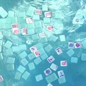 May include: A collection of light mint green mahjong tiles floating in clear blue water. The tiles feature various colorful graphic designs, including floral patterns, text, and symbols, with some tiles showing their plain backs and others showing faces.