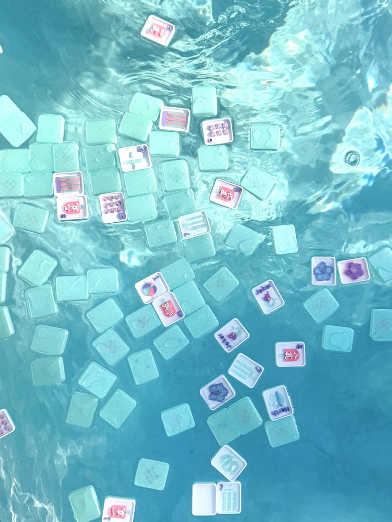 May include: A collection of light mint green mahjong tiles floating in clear blue water. The tiles feature various colorful graphic designs, including floral patterns, text, and symbols, with some tiles showing their plain backs and others showing faces.