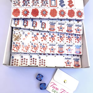 May include: A set of white mahjong tiles with red, white, and blue patriotic graphics including fireworks, roses, eagles, rockets, and top hats, alongside two blue dice and a white pouch with the text Two Tiles DESIGNS printed in pink and green.