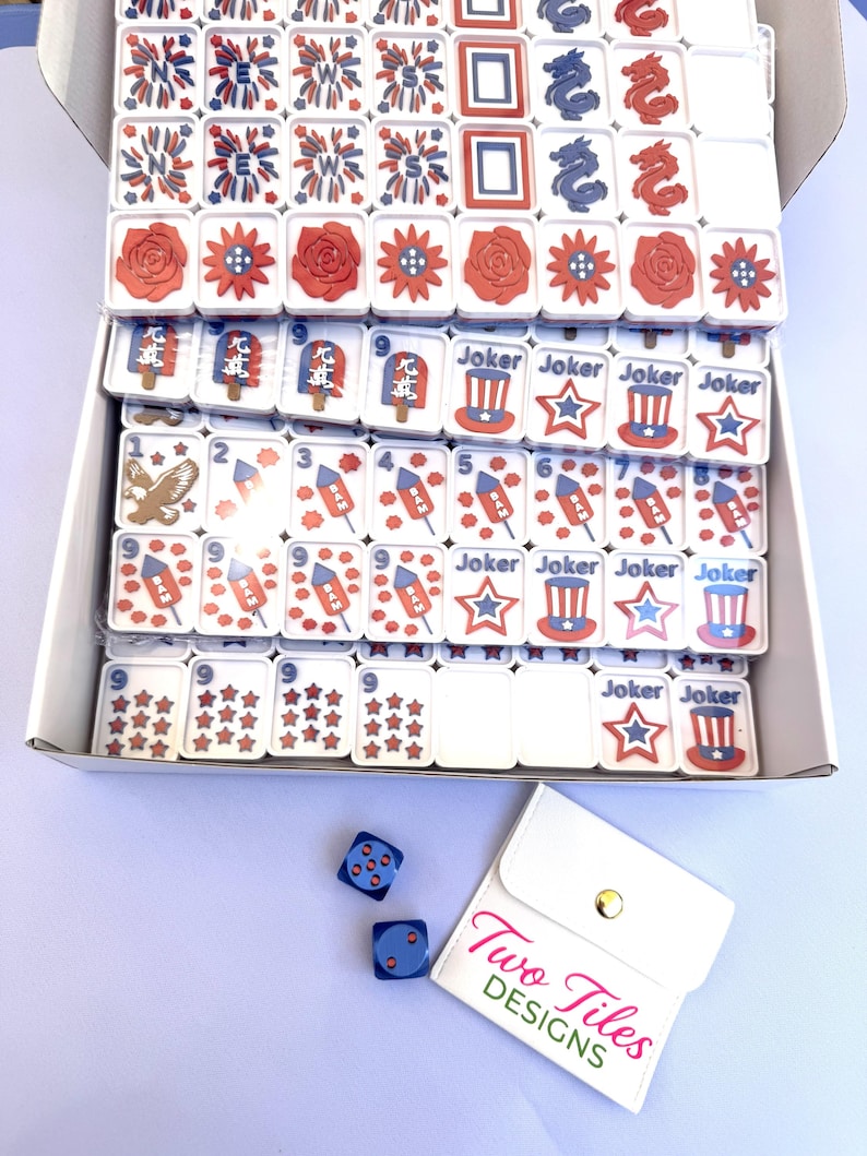 May include: A set of white mahjong tiles with red, white, and blue patriotic graphics including fireworks, roses, eagles, rockets, and top hats, alongside two blue dice and a white pouch with the text Two Tiles DESIGNS printed in pink and green.