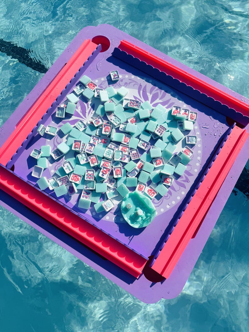 May include: A square purple and pink game board featuring a central circular area with a light purple floral pattern. Numerous small, light blue square tiles with various red and white graphic symbols are scattered across the board surface.