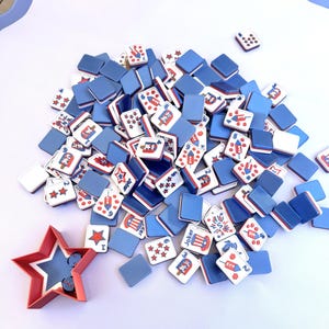 May include: A collection of rectangular game tiles featuring red, white, and blue patriotic graphics, including fireworks, stars, and top hats. A red star-shaped container holds two small blue dice. The tiles have solid blue backs and white fronts.