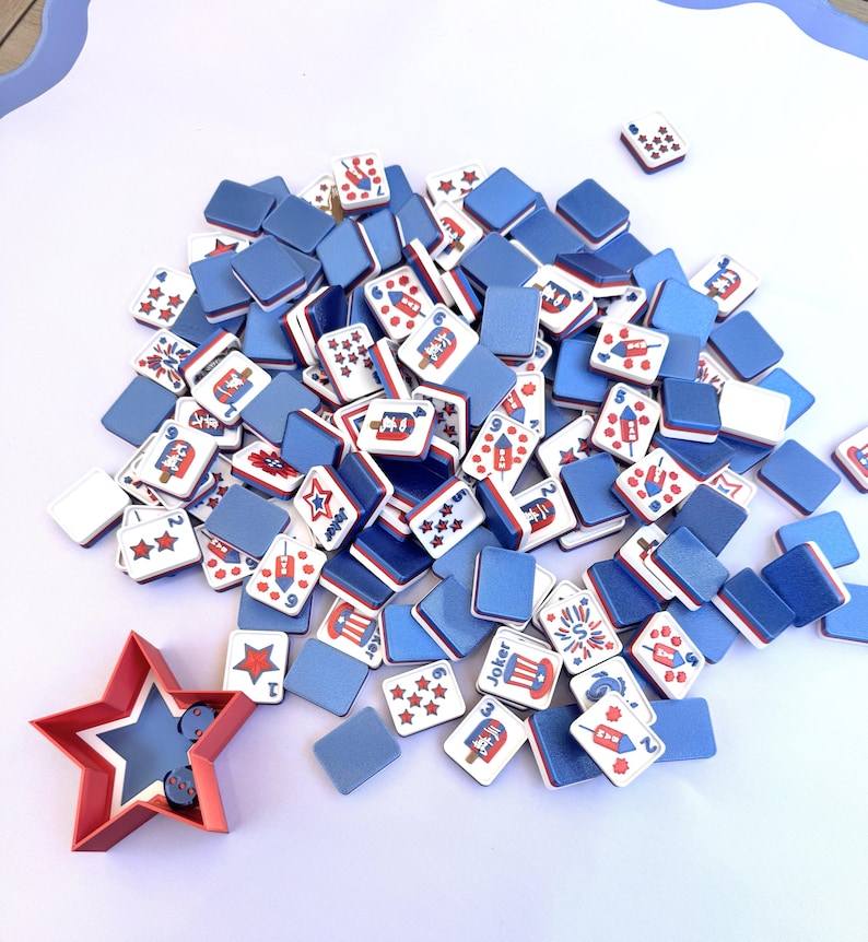 May include: A collection of rectangular game tiles featuring red, white, and blue patriotic graphics, including fireworks, stars, and top hats. A red star-shaped container holds two small blue dice. The tiles have solid blue backs and white fronts.