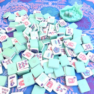 May include: A collection of light mint green mahjong tiles with white faces featuring colorful illustrations of flowers, dragons, numbers, and text like Joker, East, and West. A matching mint green shell-shaped tray holds two dice.