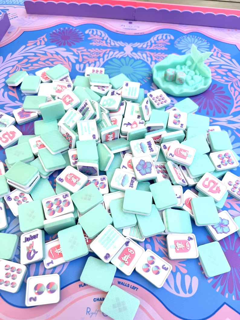 May include: A collection of light mint green mahjong tiles with white faces featuring colorful illustrations of flowers, dragons, numbers, and text like Joker, East, and West. A matching mint green shell-shaped tray holds two dice.