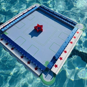 May include: A square floating game board with a light blue surface featuring green rectangular markings. The perimeter is lined with white and blue blocks decorated with red stars. A single red star-shaped piece sits in the center of the board.