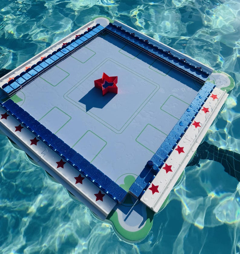 May include: A square floating game board with a light blue surface featuring green rectangular markings. The perimeter is lined with white and blue blocks decorated with red stars. A single red star-shaped piece sits in the center of the board.
